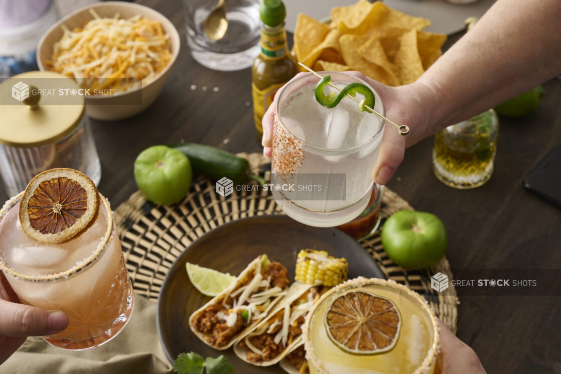 Hands Holding Assorted Citrus Margaritas Doing Cheers Over a Table of Mexican Food and Snacks in an Indoor Setting – Sequence 8