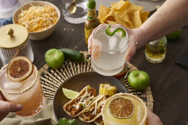 Hands Holding Assorted Citrus Margaritas Doing Cheers Over a Table of Mexican Food and Snacks in an Indoor Setting – Sequence 8