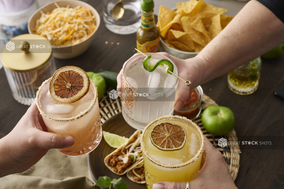 Hands Holding Assorted Citrus Margaritas Doing Cheers Over a Table of Mexican Food and Snacks in an Indoor Setting - Sequence 5