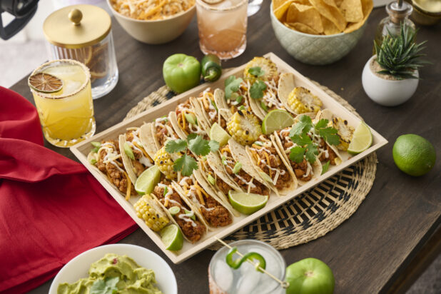 A Wood Catering Platter of Soft Tacos and Roasted Corn on a Woven Placemat, Surrounded by Citrus Margaritas on a Dark Wood Table in an Indoor Setting
