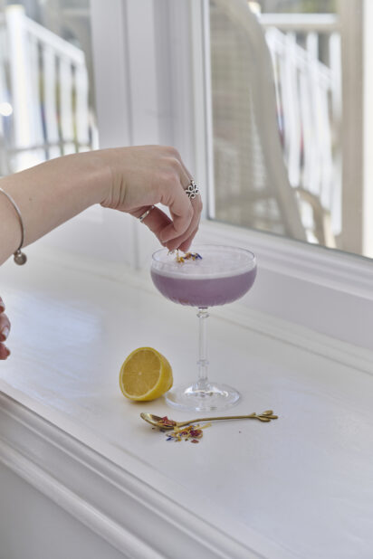 A Hand Adding Dried Flower Garnish to a Lavender Gin Sour Cocktail in a Gin Glass on a White Counter Surface with a Fresh Lemon Half in an Indoor Setting