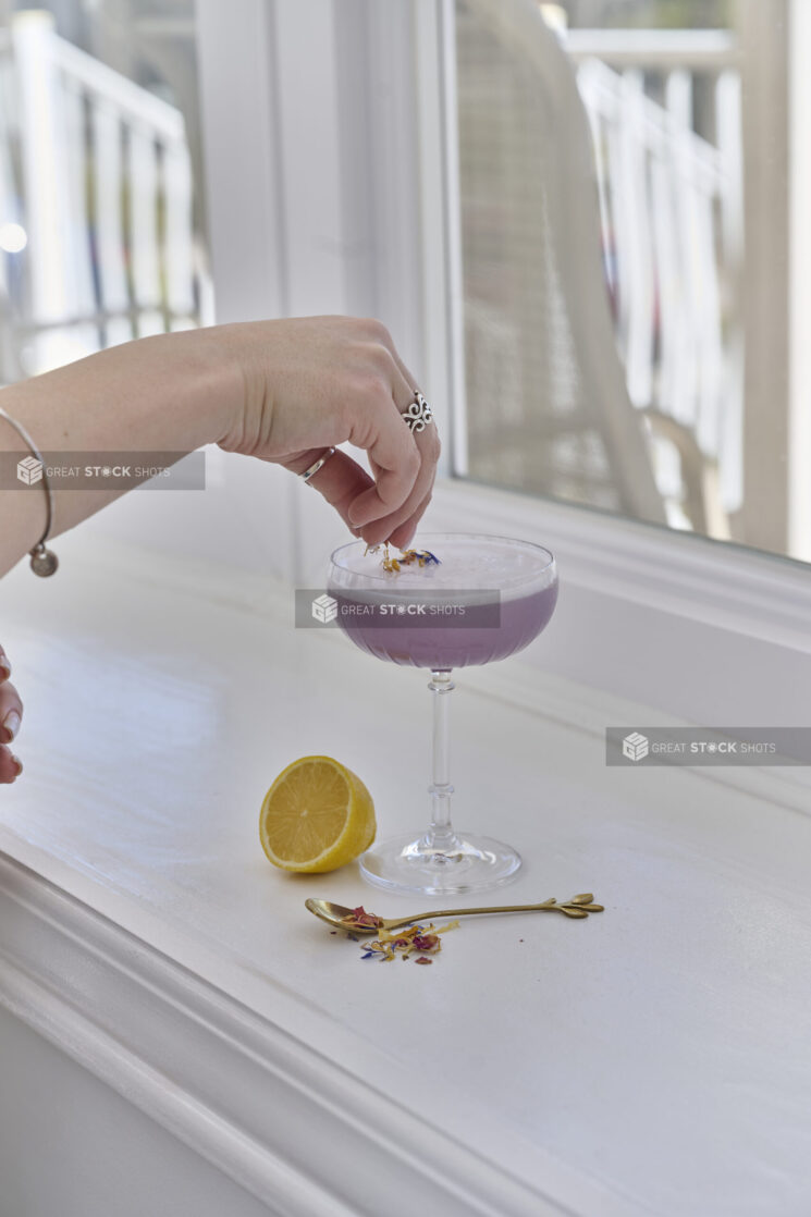 A Hand Adding Dried Flower Garnish to a Lavender Gin Sour Cocktail in a Gin Glass on a White Counter Surface with a Fresh Lemon Half in an Indoor Setting