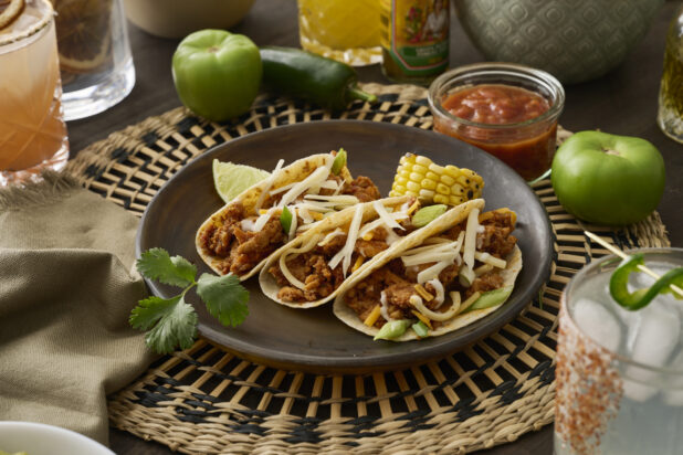 A Trio of Soft Tacos and Roasted Corn on the Cob in a Dark Brown Dish on a Woven Placemat in an Indoor Setting