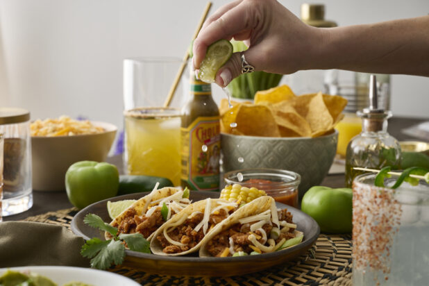 A Woman’s Hand Squeezes a Fresh Lime Wedge Over a Trio of Soft Tacos in a Dark Brown Dish in an Indoor Setting – Sequence 4