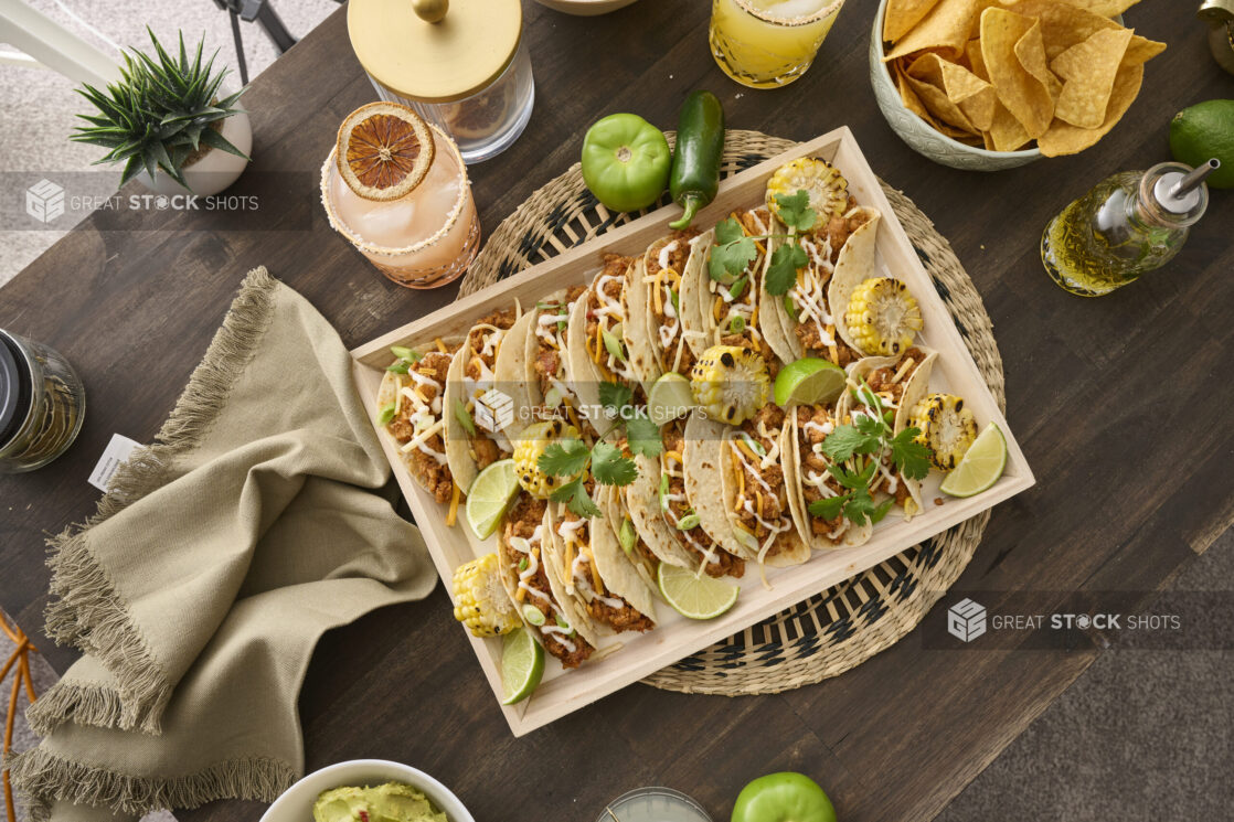 Overhead View of a Wood Catering Platter of Soft Tacos and Roasted Corn on a Woven Placemat, Surrounded by Citrus Margaritas on a Dark Wood Table in an Indoor Setting