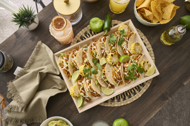 Overhead View of a Wood Catering Platter of Soft Tacos and Roasted Corn on a Woven Placemat, Surrounded by Citrus Margaritas on a Dark Wood Table in an Indoor Setting