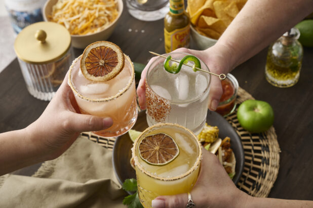 Hands Holding Assorted Citrus Margaritas Doing Cheers Over a Table of Mexican Food and Snacks in an Indoor Setting - Sequence 3