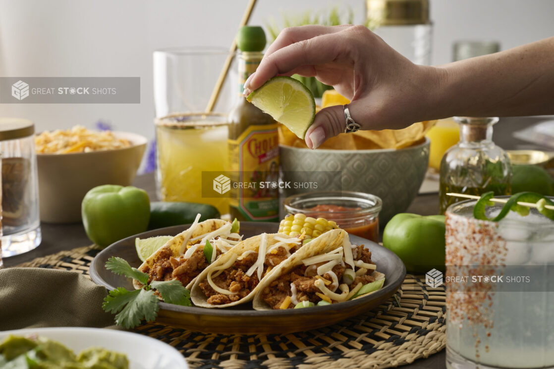 A Woman's Hand Squeezes a Fresh Lime Wedge Over a Trio of Soft Tacos in a Dark Brown Dish in an Indoor Setting - Sequence 1