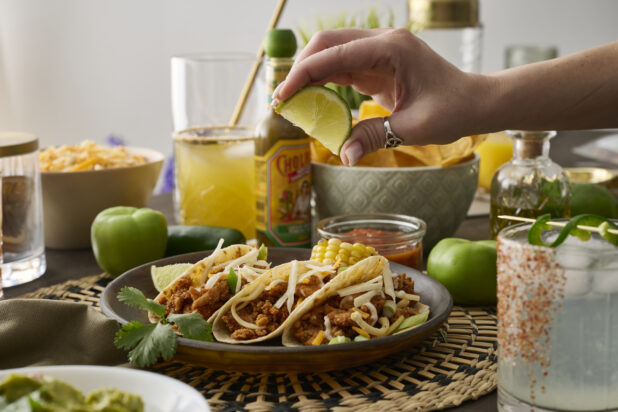 A Woman's Hand Squeezes a Fresh Lime Wedge Over a Trio of Soft Tacos in a Dark Brown Dish in an Indoor Setting - Sequence 1