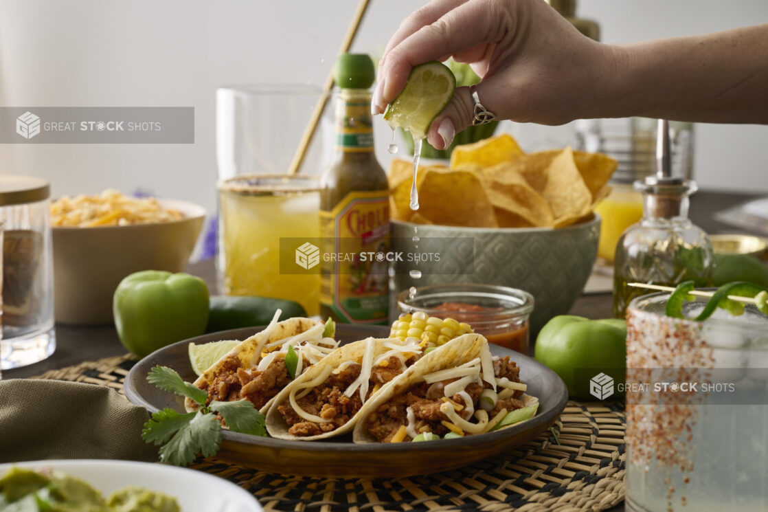 A Woman’s Hand Squeezes a Fresh Lime Wedge Over a Trio of Soft Tacos in a Dark Brown Dish in an Indoor Setting – Sequence 3