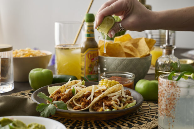 A Woman’s Hand Squeezes a Fresh Lime Wedge Over a Trio of Soft Tacos in a Dark Brown Dish in an Indoor Setting – Sequence 3
