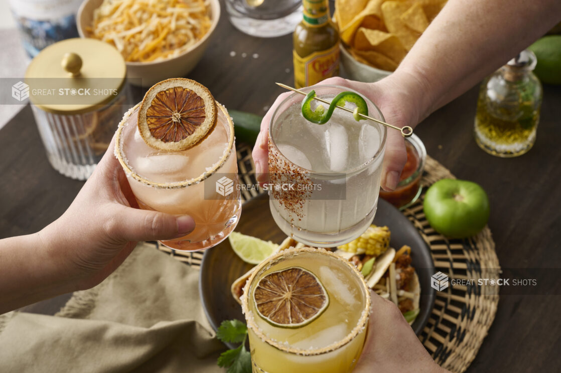 Hands Holding Assorted Citrus Margaritas Doing Cheers Over a Table of Mexican Food and Snacks in an Indoor Setting - Sequence 4