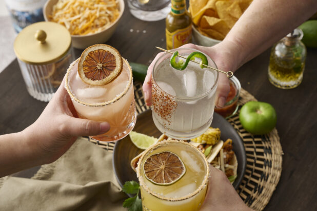Hands Holding Assorted Citrus Margaritas Doing Cheers Over a Table of Mexican Food and Snacks in an Indoor Setting - Sequence 4