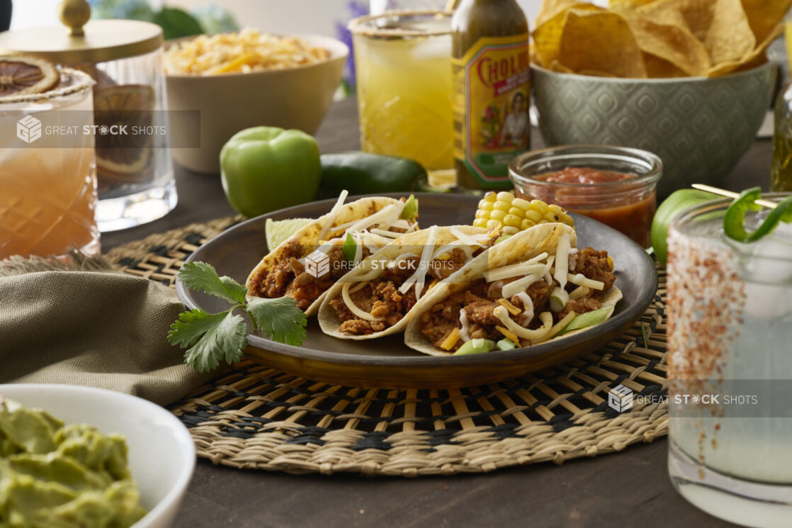 Close-Up of a Trio of Soft Tacos and Roasted Corn on the Cob in a Dark Brown Dish on a Woven Placemat in an Indoor Setting