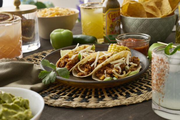 Close-Up of a Trio of Soft Tacos and Roasted Corn on the Cob in a Dark Brown Dish on a Woven Placemat in an Indoor Setting