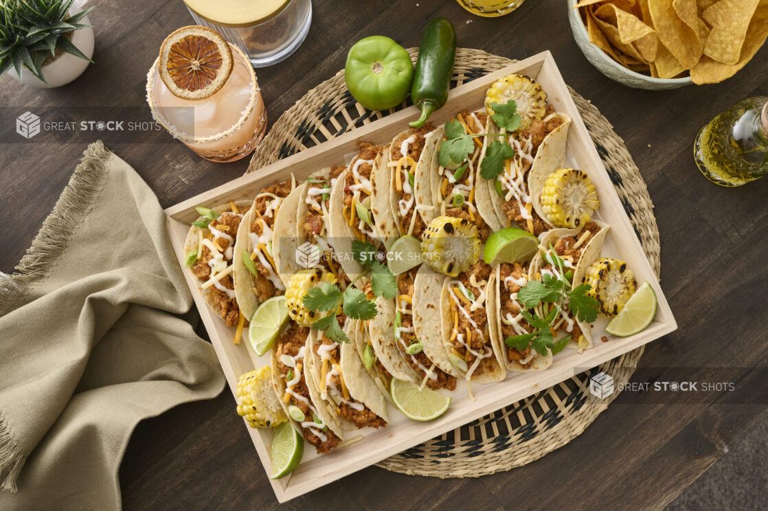 Overhead View of a Wood Catering Platter of Soft Tacos and Roasted Corn on a Woven Placemat, Surrounded by Citrus Margaritas on a Dark Wood Table in an Indoor Setting - Variation